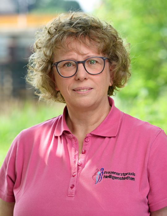 Foto von Angelika Hempel, sie lächelt, hat gelockte blonde Haare, trägt eine Brille und ein pinkes Shirt mit dem Logo der Praxis.