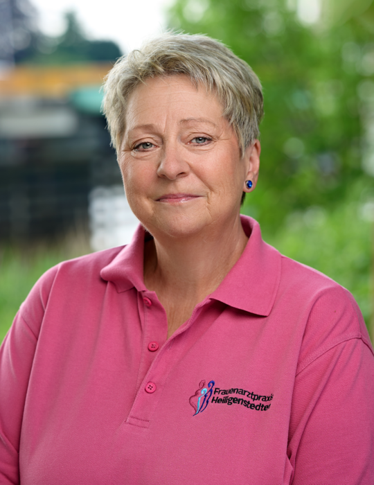 Foto von Heike Busch, sie lächelt, hat blonde, kurze Haare und trägt ein pinkes Shirt mit dem Logo der Praxis.